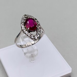 Könnte beinhalten: Ein silberner Ring mit einem großen ovalen Rubin, der von einem Heiligenschein aus kleineren weißen Edelsteinen umgeben ist.