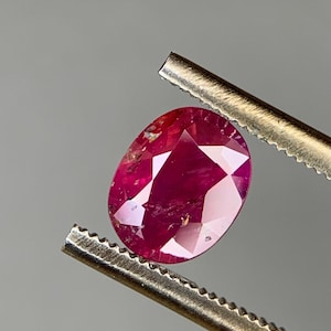 Peut inclure: Une pierre précieuse rubis ovale et facettée, tenue par une pince. Le rubis est d'un rouge profond avec des inclusions visibles et des facettes qui captent la lumière. La pince est de couleur argentée, sur fond gris.