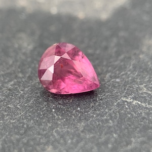 Op de afbeelding: Een peervormige roze edelsteen met een facetgeslepen vorm. De edelsteen is geplaatst tegen een donkergrijze achtergrond.