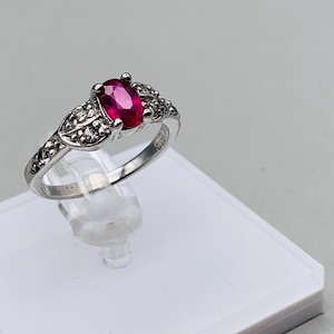 Könnte beinhalten: Ein silberner Ring mit einem roten ovalen Edelstein und kleineren klaren Edelsteinen auf der Ringschiene. Der Ring befindet sich auf einem weißen Präsentationsständer.