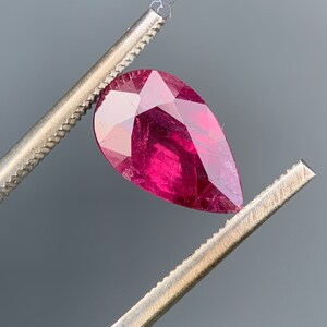 May include: A pear-shaped ruby gemstone held by tweezers. The ruby is a deep red colour and has a faceted cut.