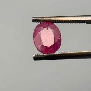 May include: A vibrant pink oval-shaped gemstone held by tweezers. The gemstone has a faceted cut and is translucent.