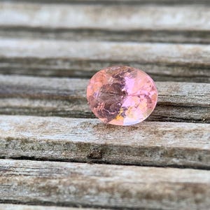 Könnte beinhalten: Ein facettierter ovaler Edelstein mit einem durchscheinenden hellrosa Farbton. Der Stein weist einen Brillantschliff mit mehreren dreieckigen und rautenförmigen Facetten auf, die das Licht reflektieren und eine funkelnde Oberfläche erzeugen.
