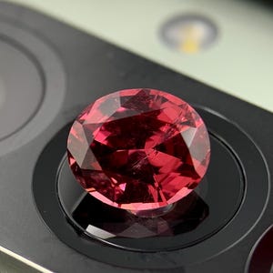 Könnte beinhalten: Ein facettierter, runder, tiefroter Edelstein. Der Edelstein ist auf einer schwarzen Oberfläche mit kreisförmigen Details angeordnet. Die Facetten des Edelsteins reflektieren das Licht und erzeugen einen funkelnden Effekt. Der Hintergrund ist eine dunkle, reflektierende Oberfläche.
