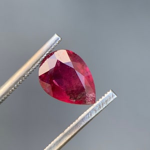 May include: A pear-shaped, deep red gemstone held by tweezers. The gemstone has a faceted cut and is translucent.