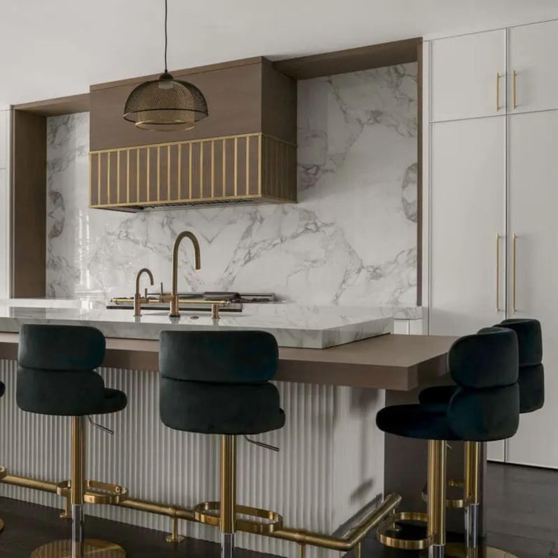 May include: A modern kitchen with a marble backsplash and countertop. Features include a brown range hood with gold accents, gold faucets, and black velvet bar stools with gold bases. A pendant light hangs above the island.