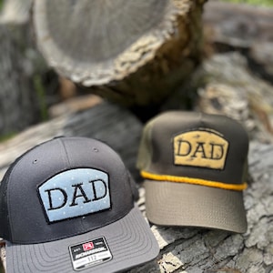 Op de afbeelding: Twee baseballpetten met het woord "DAD" op de voorkant. Eén pet is grijs met een zwarte mesh achterkant en een lichtblauwe patch met witte stippen. De andere is olijfgroen met een bruine patch en een gele touwdetail.