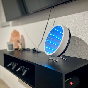 May include: A white round mirror with a blue LED light display on a black shelf. The mirror is on a metal stand.