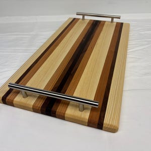 Op de afbeelding: Een rechthoekig houten serveerblad met afwisselende verticale strepen van licht-, midden- en donkerbruine houtnerven. Het blad is voorzien van twee horizontale handgrepen van geborsteld metaal aan de kortere zijden van het bord.