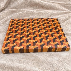 May include: A rectangular wooden cutting board featuring an intricate geometric pattern composed of light, medium, and dark brown wood segments. The design creates a three-dimensional cube effect across the surface, with a smooth, flat top.