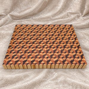 Puede incluir: Una tabla de cortar rectangular de madera con un intrincado patrón geométrico de cubos entrelazados en varios tonos de madera clara, media y oscura. La superficie muestra un diseño repetitivo de cubos en tres dimensiones.