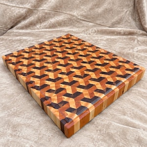May include: A rectangular wooden cutting board featuring an intricate geometric pattern composed of light, medium, and dark brown wood segments. The surface displays a repeating three-dimensional cube design with smooth, polished edges and sides.