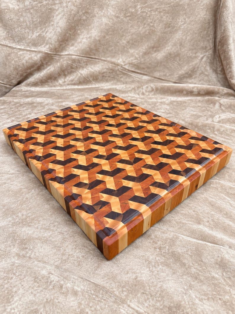 May include: A rectangular wooden cutting board featuring an intricate geometric pattern composed of light, medium, and dark brown wood segments. The surface displays a repeating three-dimensional cube design with smooth, polished edges and sides.