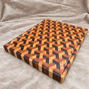 May include: A rectangular wooden cutting board featuring an intricate geometric pattern composed of light, medium, and dark brown wood segments. The surface displays a repeating three-dimensional cube design with a smooth, polished finish.
