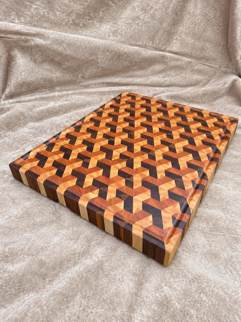 May include: A rectangular wooden cutting board featuring an intricate geometric pattern composed of light, medium, and dark brown wood segments. The surface displays a repeating three-dimensional cube design with a smooth, polished finish.