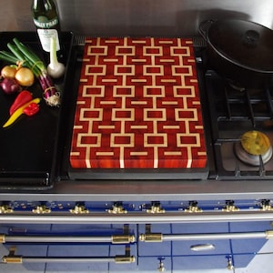 Puede incluir: Una tabla de cortar rectangular de madera con un patrón geométrico de bloques de madera de color rojo oscuro y beige claro. La tabla está colocada sobre una estufa azul con mangos dorados, junto a una olla negra, una botella y verduras frescas.