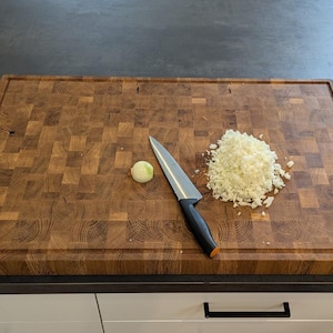 Puede incluir: Una tabla de cortar de madera con un patrón de cuadros, una cebolla blanca, un cuchillo y cebolla blanca picada.