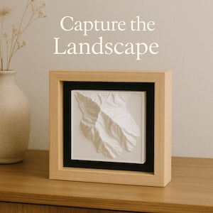 May include: A framed, three-dimensional white relief map of a mountain range. The artwork is set within a black mat and a light wood frame. The text "Capture the Landscape" is above the frame.