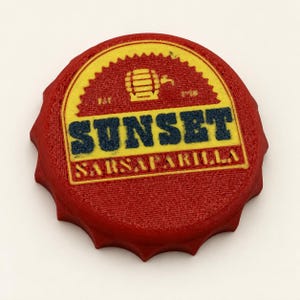 May include: A circular red bottle cap with a yellow centre featuring a graphic of a barrel and the text SUNSET SARSAPARILLA with EST 1918 printed on the sides. The design has a textured surface and a scalloped edge typical of a bottle cap.