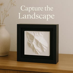 Könnte beinhalten: Ein schwarz gerahmtes 3D-Reliefbild eines Gebirges mit weißen, strukturierten Gipfeln vor weißem Hintergrund. Der Text "Capture the Landscape" befindet sich über dem Rahmen. Das Kunstwerk wird auf einer Holzoberfläche präsentiert.