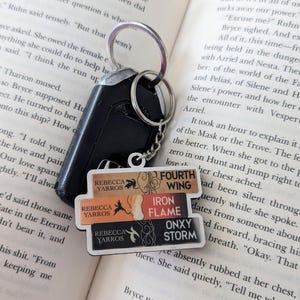 Könnte beinhalten: Nahaufnahme eines Buches mit einem daran befestigten Schlüsselanhänger. Der Schlüsselanhänger hat drei buchförmige Anhänger mit den Titeln "Fourth Wing", "Iron Flame" und "Onyx Storm". Die Buchtitel sind in weißer Schrift auf schwarzem Hintergrund. Die Anhänger sind übereinander gestapelt. Das Buch ist auf einer Seite mit Text über die Charaktere Azriel und Nesta aufgeschlagen.