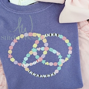 May include: Purple t-shirt featuring a colourful beaded design that spells out "Fearless" and "1989" in black text.
