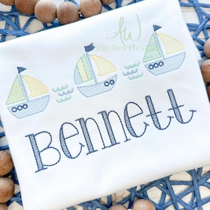 May include: A white onesie with blue and yellow embroidered sailboats and the name "Bennett" in blue embroidery.