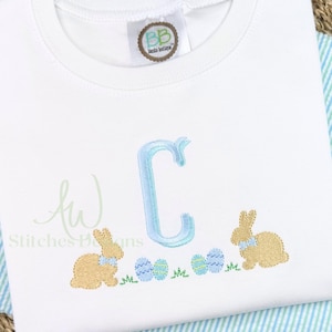 May include: A white t-shirt with a blue embroidered letter C, two brown embroidered bunnies, and green and blue embroidered Easter eggs. The text "Aw Stitches Designs" is embroidered in the bottom left corner.