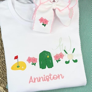 May include: White embroidered baby onesie with a green golf jacket, golf clubs, golf ball, and pink flowers. The onesie is personalized with the name "Anniston".
