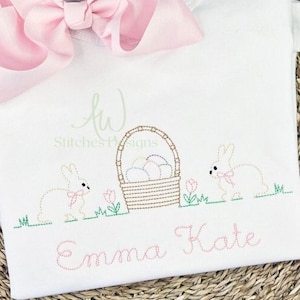 Puede incluir: Una camisa blanca bordada con dos conejos, una cesta de huevos de Pascua y el nombre "Emma Kate" en hilo rosa.