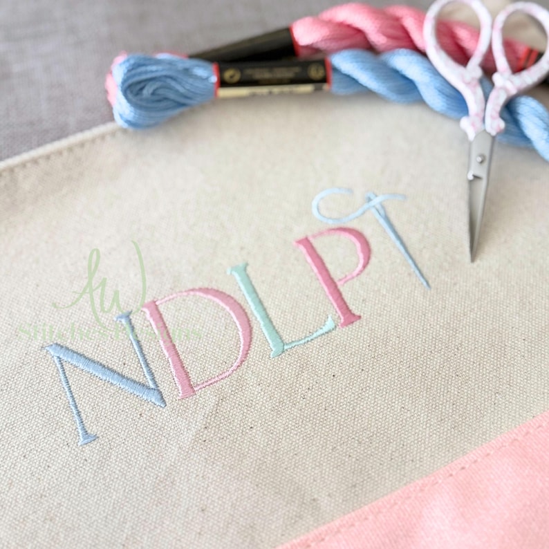 May include: A canvas tote bag with embroidered initials in pastel colors. The letters "NDLP" are stitched in light blue and pink thread. A pair of small scissors and spools of thread in matching colors are also visible.