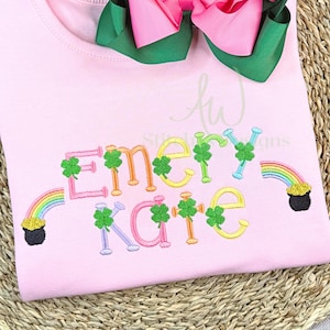 May include: Pink t-shirt with embroidered text "Emery Kate" in colorful letters, decorated with shamrocks, rainbows, and a pot of gold. A large pink and green bow is at the top. The shirt is likely for a child.