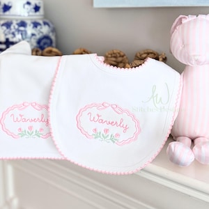 May include: White baby bib and burp cloth set with pink scalloped edges. Each item features an embroidered pink oval with the name "Waverly" and floral accents. A pink and white striped stuffed animal is in the background.