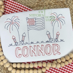 May include: A white embroidered shirt with a red, white, and blue American flag, fireworks, and the name "CONNOR" in red.