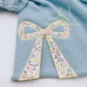 May include: A light blue knitted sweater with a floral patterned bow applique. The bow is outlined in yellow.