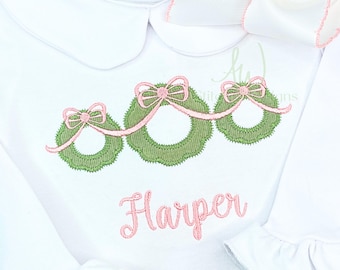 Op de afbeelding: Witte baby romper met een geborduurd ontwerp in roze en groen van drie kransen met strikken en de naam "Harper" eronder.