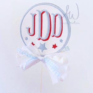 May include: A white embroidered circle with the letters "JDD" in red and blue, surrounded by a silver border and stars. A white bow with blue gingham accents is attached to a wooden stick. The design is patriotic.