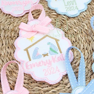 May include: Embroidered Christmas ornament with a pink scalloped edge, featuring a nativity scene with Mary, Joseph, and baby Jesus. The ornament has a pink ribbon bow and the name "Emery Kat" and the year "2024" embroidered in pink.