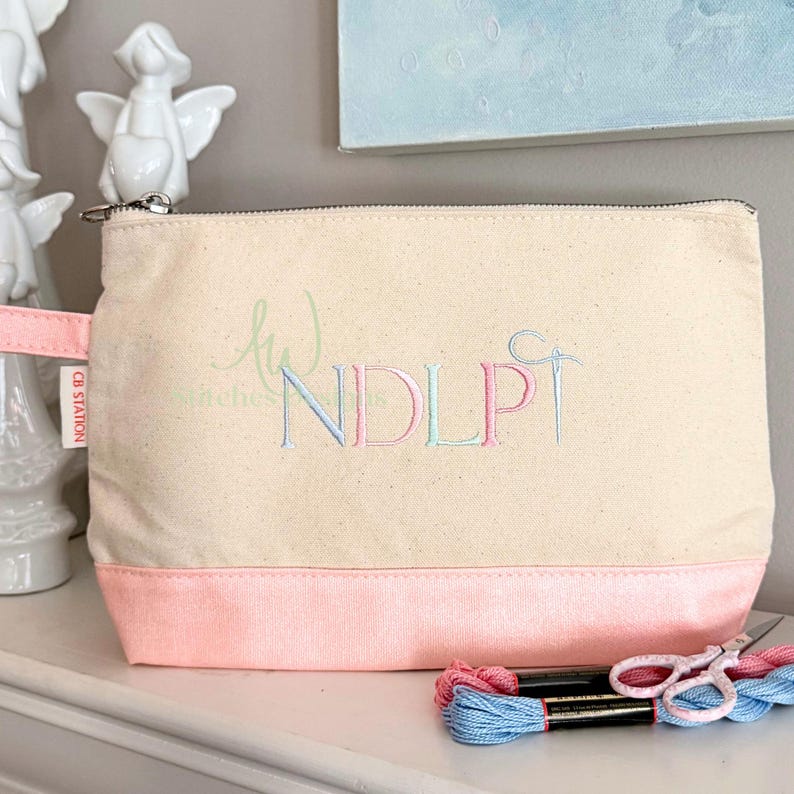 May include: A canvas zippered pouch with a pink base and handle. The pouch features embroidered letters "NDLP" in pastel colors, with a needle and thread design. Embroidery floss and scissors are visible.