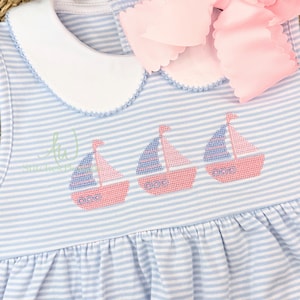 Op de afbeelding: Blauw en wit gestreepte babyjurk met geborduurde zeilboten. De jurk heeft een witte kraag, een roze strik en drie geborduurde zeilboten met roze rompen en blauwe zeilen. De jurk heeft een gerimpelde rok.