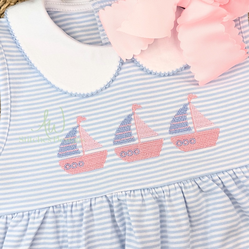 Op de afbeelding: Blauw en wit gestreepte babyjurk met geborduurde zeilboten. De jurk heeft een witte kraag, een roze strik en drie geborduurde zeilboten met roze rompen en blauwe zeilen. De jurk heeft een gerimpelde rok.