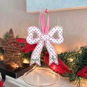 May include: A white bow-shaped Christmas ornament with red and green polka dots. The bow has a pink ribbon for hanging and features the year "2025" embroidered in green. The ornament is displayed on a clear stand.