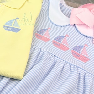 Op de afbeelding: Een gele poloshirt en een blauw-wit gestreepte jurk. Beide kledingstukken hebben geborduurde zeilbootontwerpen in blauw en roze. De jurk heeft een witte kraag en een roze strik. Kinderkleding.