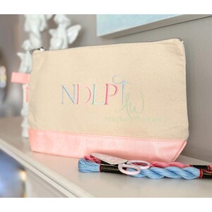 May include: A beige canvas zippered pouch with a pink bottom panel and a pink fabric loop. The pouch has embroidered text that reads "NDLP" and "kw Stitches & Designs" in pastel colors. A bundle of blue and pink embroidery floss and small scissors are in front.