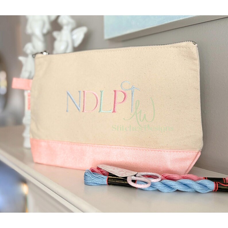 May include: A beige canvas zippered pouch with a pink bottom panel and a pink fabric loop. The pouch has embroidered text that reads "NDLP" and "kw Stitches & Designs" in pastel colors. A bundle of blue and pink embroidery floss and small scissors are in front.