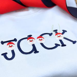 May include: A light blue t-shirt with the name "JACK" embroidered in navy blue with a Santa Claus face in each letter.