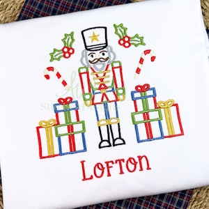 May include: White t-shirt with an embroidered Christmas design. The design features a nutcracker soldier, candy canes, holly, and wrapped gifts. The name "Lofton" is embroidered in red script below the design.