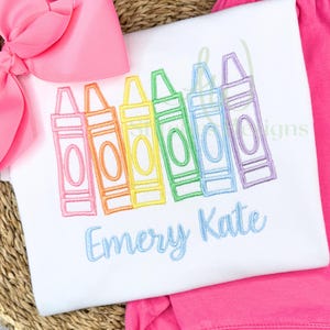 Embroidered design of colorful crayons with the name 'Emery Kate' on a white fabric background.