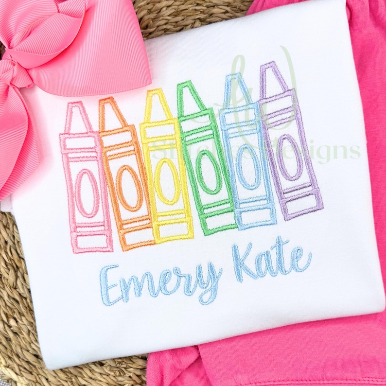 Embroidered design of colorful crayons with the name 'Emery Kate' on a white fabric background.