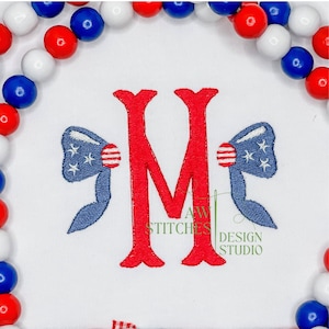 May include: Red, white, and blue embroidered letter M with two bows on either side. The bows are blue with white stars and red stripes. The text "AW STITCHES DESIGN STUDIO" is below the letter.
